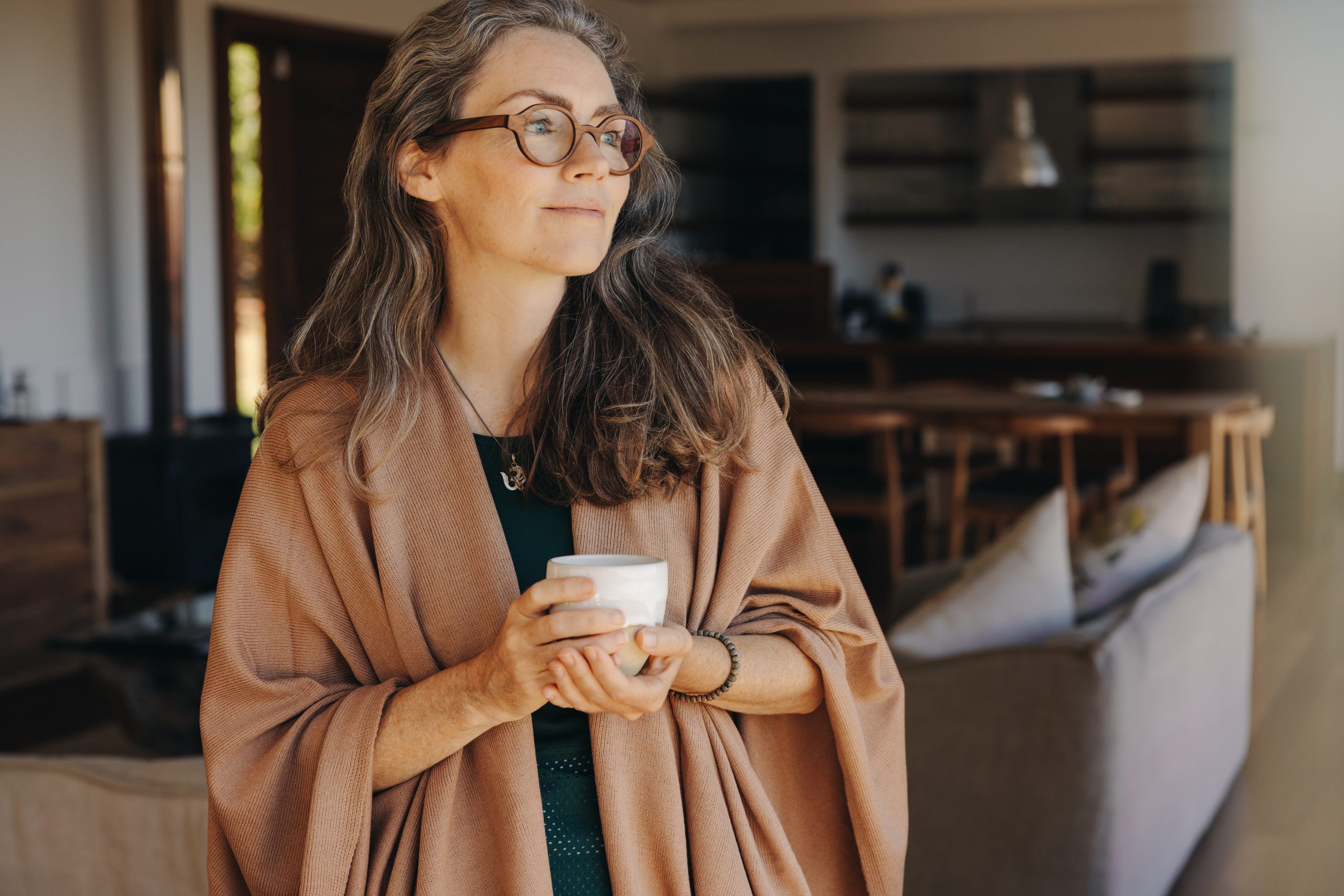 Thoughtful senior woman holding a tea cup at home Senior woman looking away thoughtfully while standing with a cup of tea in her hands. Mature woman enjoying a serene retirement at home.