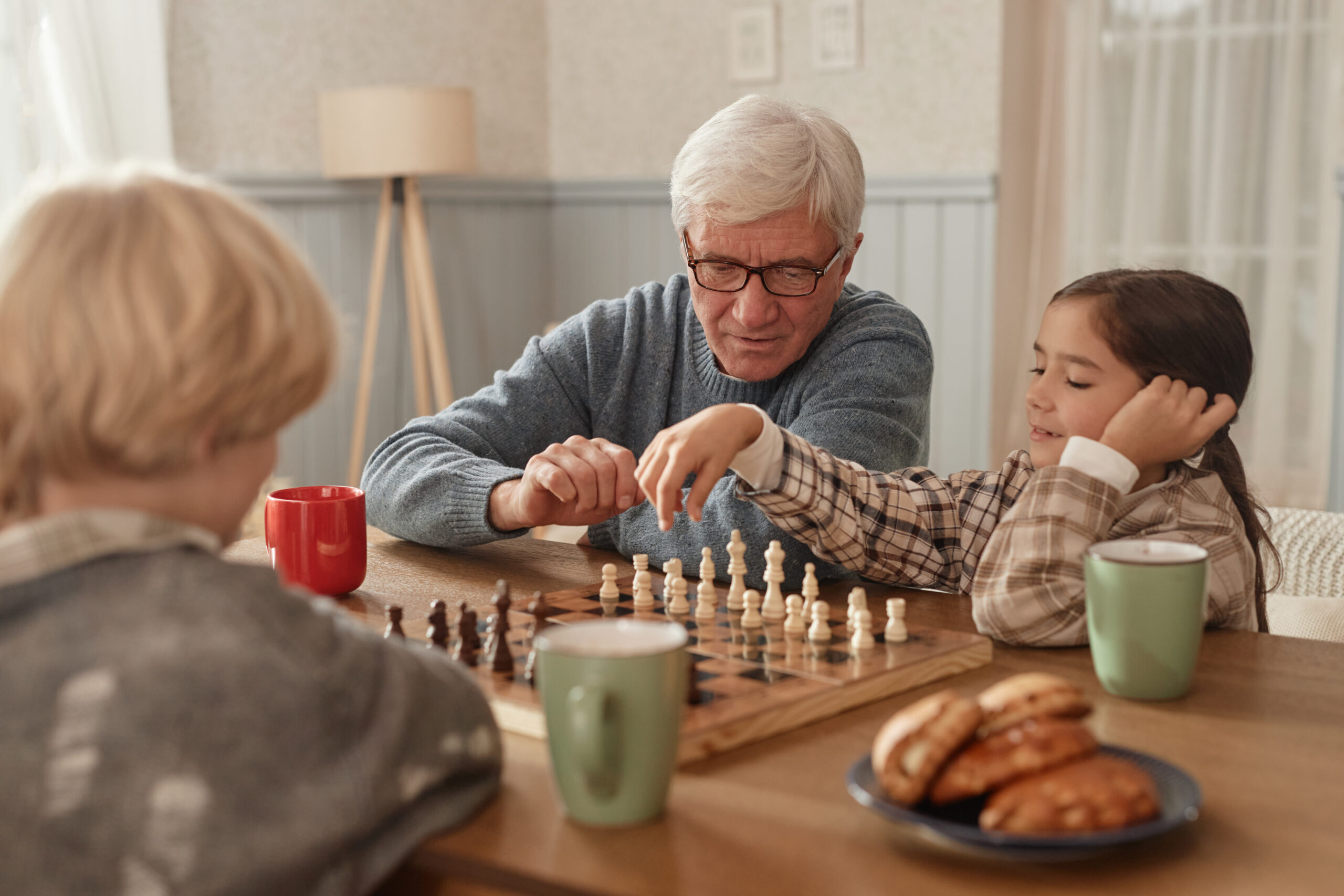 Senior Caucasian man teaching chess to boy and girl at wooden table in home, children concentrating on game, mugs and pastries visible in foreground