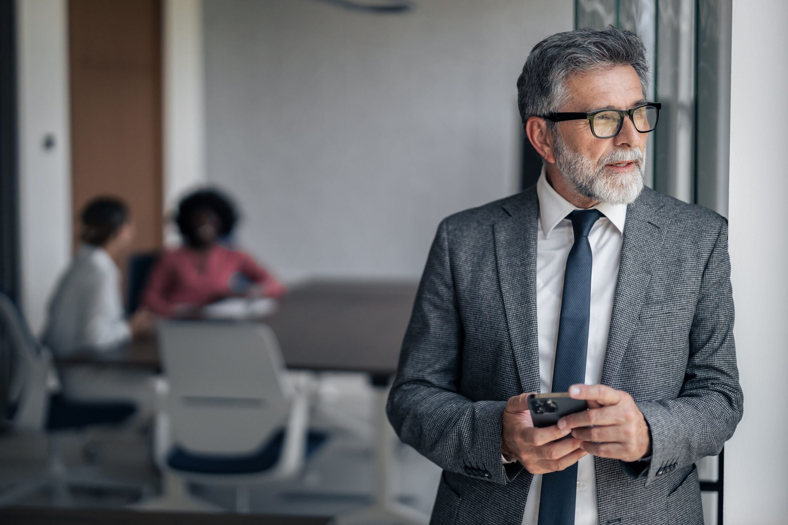 Experienced man in a formal suit and eyeglasses holding a smartphone, looking thoughtful, with blurred colleagues discussing in the background of a modern office environment