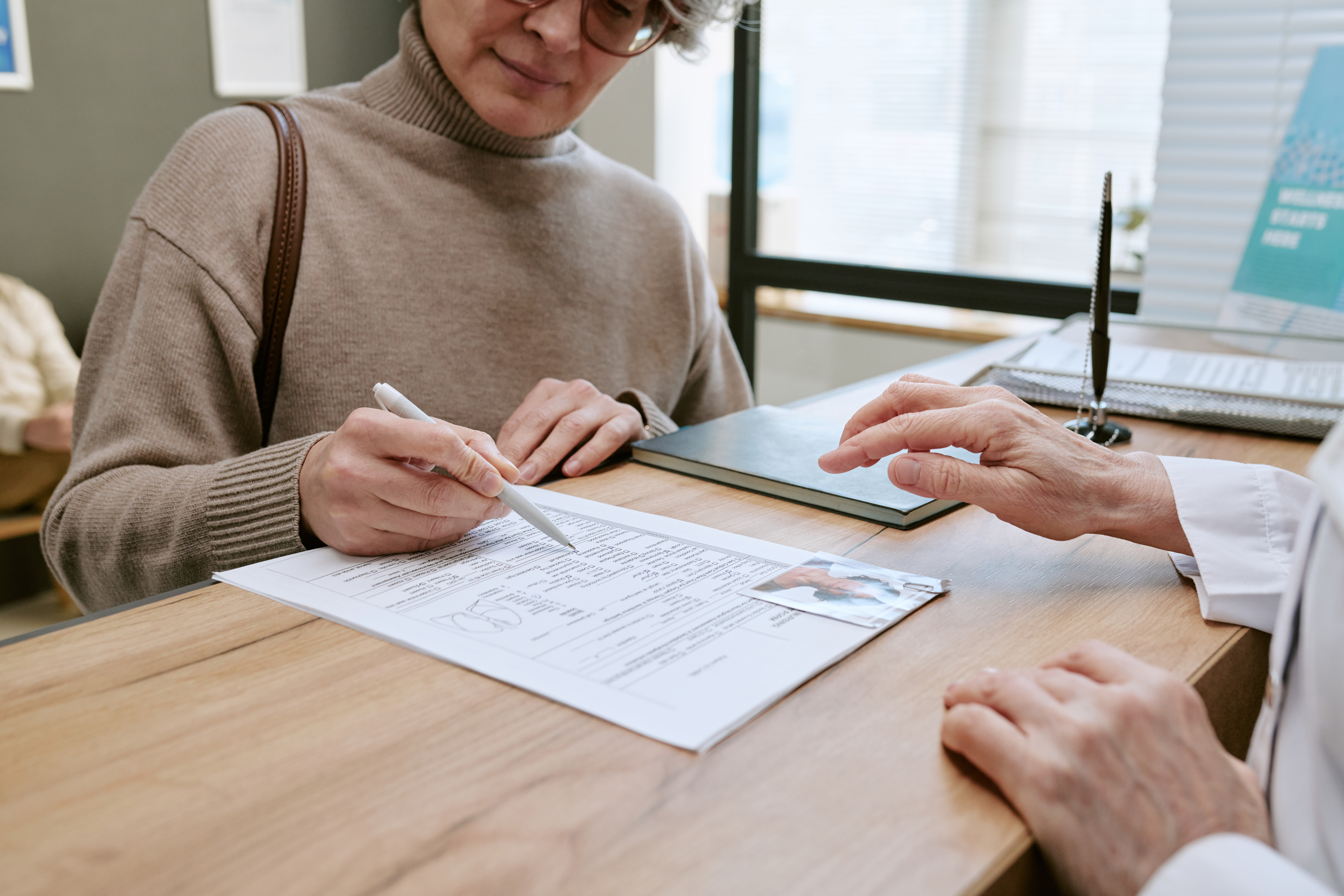 Middle aged Caucasian woman signing medical form while doctor guiding paperwork at clinic desk, representing patient intake and healthcare administration use in insurance or treatment marketing