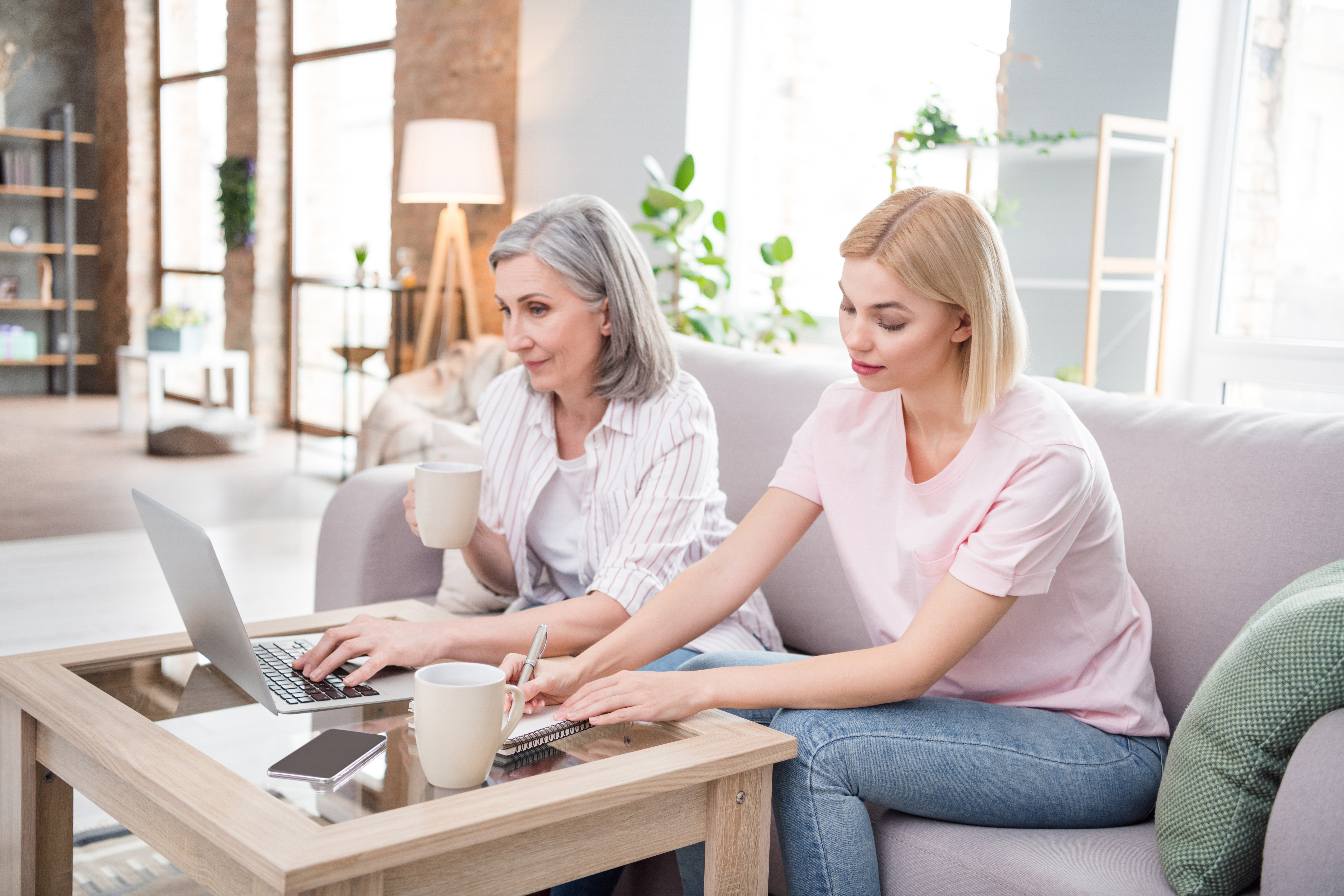 Photo of serious family grey haired mom and blonde daughter sit sofa write note use laptop indoors inside house home.